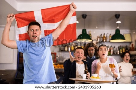 Company of young adult sports fans supporting Danish team with state flag while drinking beer in bar
