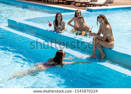 Company of young merry girls and guy relaxing in the swimming pool drinking cocktails on the open air on a sunny summer day next to lounge zone with sun lounger