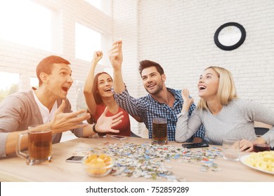 The Company Of Young People Is Folding The Image. On The Table They Have Puzzles And Stand Glasses With Alcoholic Beverages. They Have Fun Playing A Game.