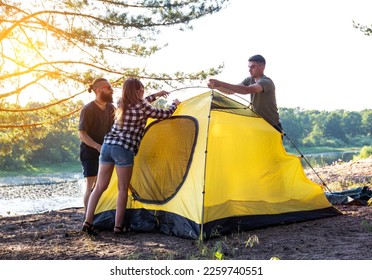 A company of young people, girls and guys, set up a tent in nature. Outdoor recreation in summer. Beautiful sunny day on the banks of the river and forest, tourism. - Powered by Shutterstock