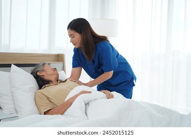 Compassionate nurse assistant offering gentle care to senior woman lying in bed. Young caregiver ensures elderly lady's comfort and warmth, covering her with soft blanket for restful sleep at home. - Powered by Shutterstock