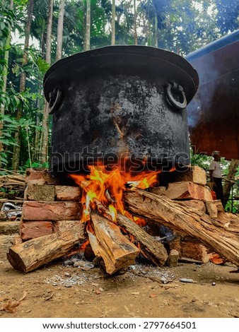 ​A compelling low angle, vertical photograph capturing the essence of traditional community cooking in a rural setting. The center of the frame features a massive, soot covered black iron cauldron with ring handles resting on a makeshift stove built from red clay bricks. Below the pot, bright orange and yellow flames erupt from a roaring wood fire fueled by thick, rustic logs. The foreground shows scattered ash on the dirt ground, while the background reveals a lush tropical environment filled with tall betel nut or palm trees and a partial view of a shelter. In the soft-focus background on the right, an elderly village man stands quietly, adding an authentic, human storytelling element to this cultural culinary scene. This highly vivid image is perfect for cultural documentaries, food heritage blogs, and regional tourism publications.