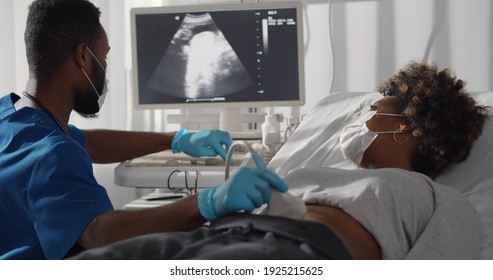 Competent Afro Male Doctor In Medical Protective Mask Doing Ultrasound Screening For Pregnant Woman At Modern Clinic. Young Afro Woman In Safety Mask Visiting Gynecologist