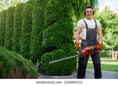 Competent caucasian gardener smiling and looking at camera while standing on back yard and holding hand electric trimmer. Man using gardening tool for cutting hedge.  - Powered by Shutterstock