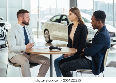 Competent Salesman Talking With Multiracial Couple About Purchasing Of New Car. Caucasian Woman And African Man Buying Vehicle At Showroom. Selling And Business Concept.