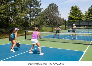 A competitive doubles game of pickleball at the net on a blue and green court in summer.