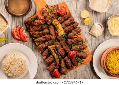 A complete Middle Eastern meal featuring grilled beef kofta served on a rustic wooden board. The platter is accompanied by traditional Egyptian rice with vermicelli, yellow basmati rice, a bowl of warm soup, creamy tahini, and fresh slices of onion, tomato, and lemon. Captured in a professional top-view (flat lay) style on a light wooden background.