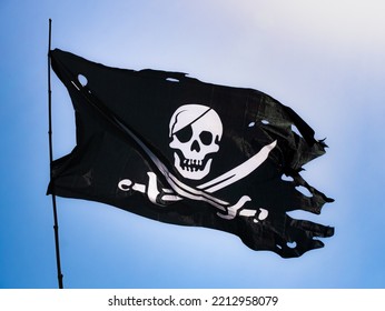 Completely torn and bruised black colored pirate flag (Jolly Roger) fastened to a flagpole flutters in the wind with a skull with patch and two swords on the bottom in white drawn on the fabric - Shutterstock ID 2212958079