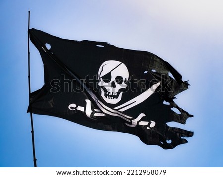 Completely torn and bruised black colored pirate flag (Jolly Roger) fastened to a flagpole flutters in the wind with a skull with patch and two swords on the bottom in white drawn on the fabric
