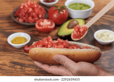 Completo Italian. Chilean: Adding tomato, with a wooden spoon, on a hot dog bun, held with one hand, tomato, avocado, mayonnaise and mustard on a wooden table. Typical Chilean food concept - Powered by Shutterstock