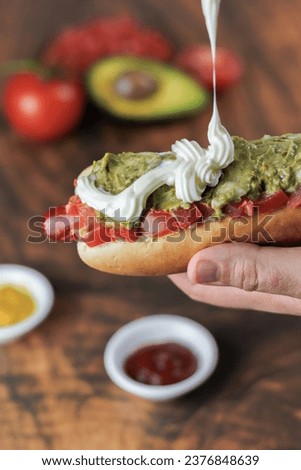 completo italiano: Close-up vertical. Adding mayonnaise, over tomato and vianesa on hot dog bun, held with one hand, tomato, palta and mustard on a wooden table. Typical Chilean food concept