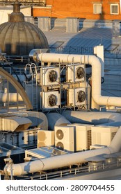 Complex industrial HVAC setup on flat roof. Multiple white AC units stacked in metal frames. Thick white pipes weave across structure. Warm sunset light highlights metallic surfaces. air conditioning.