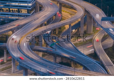 A complex network of elevated highways curves through a modern city, illuminated by streetlights and streaks of moving vehicle lights at dusk.