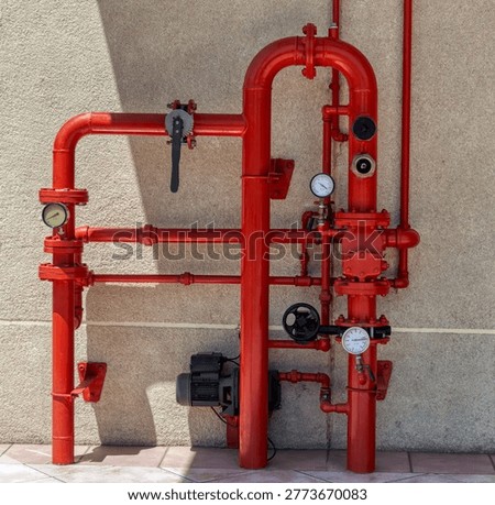 complex network of red painted metal pipes, valves, and pressure gauges for a fire protection system is mounted on an exterior textured wall.