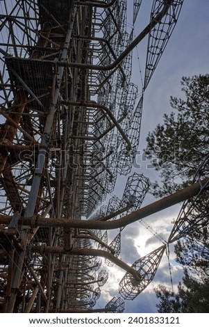 A complex structure of rusted metal beams and wire grids against a cloudy sky, surrounded by trees. Duga is a Soviet over-the-horizon radar station for an early detection system for ICBM launches.