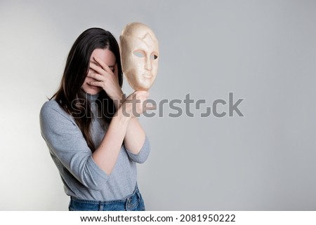 Complexes due to appearance or self-identification problems, concept. A young woman holds a mask in her hands hiding her face. Photos in light gray tones, gray background