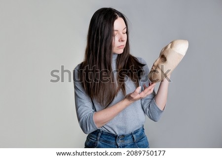 Complexes due to appearance or self-identification problems, concept. A young woman holds a mask in her hands hiding her face. Photos in light gray tones, gray background