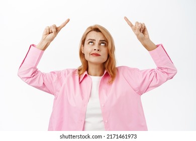 Complicated Girl Looks Uneasy And Points Up, Showing Smth On Top With Perplexed Face Expression, Standing Over White Background