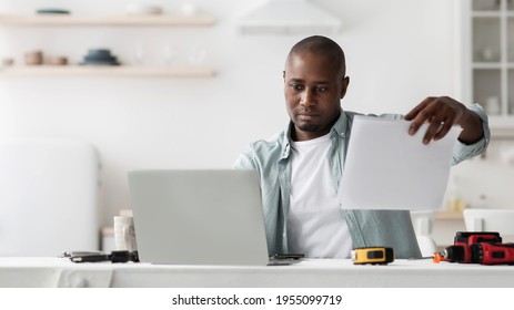 Complicated Instructions. Serious Black Handyman At Desk With Laptop And Tools For Repair Reading Specification And Comparing With Online Manual, Sitting In Kitchen, Panorama With Empty Space