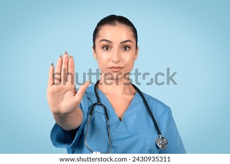 Composed female medical professional in blue scrubs makes firm stop gesture, conveying authority and importance of health protocols against serene blue backdrop