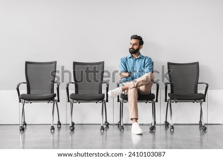 Composed indian businessman seated with legs crossed, reflecting alone in an empty row of chairs in a sparse and modern waiting area, full length