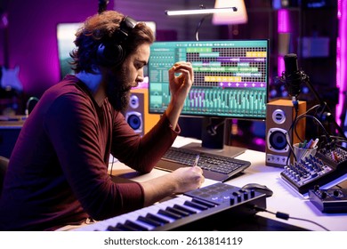 Composer songwriter taking notes to create a new song in his home studio, writing down lyrics and musical notes before recording his tune on mixing console and microphone, music industry.