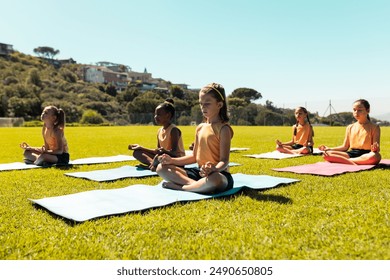 Composite of diverse schoolgirls meditating in lotus pose while sitting on mats over grassy field. Copy space, yoga, zen, exercise, fitness, childhood, nature, summer, together, german, school. - Powered by Shutterstock