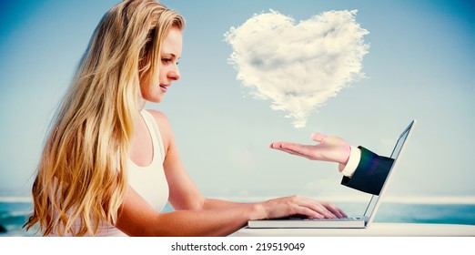 Composite image of pretty blonde using her laptop at the beach with hand presenting cloud heart