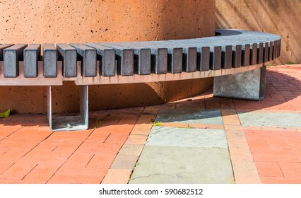 Composite And Steel Semicircular Public Bench Set Against An Orange Painted Concrete Column And Casting Shadows On Brick And Slate Pavers.