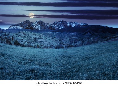 Composite Summer Landscape With 3D Mountains. Village On Agricultural Meadow Hillside In Front Of A High Rocky Mountain Range At Night In Full Moon Light