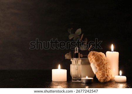 Composition with bath sponge, bottle of cosmetic product and burning candles on black background, closeup