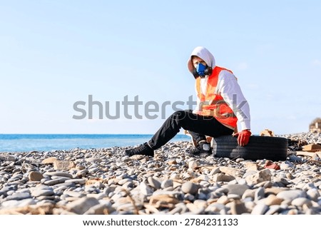 Composition capturing masked volunteer inspecting tire among beach waste under clear weather.