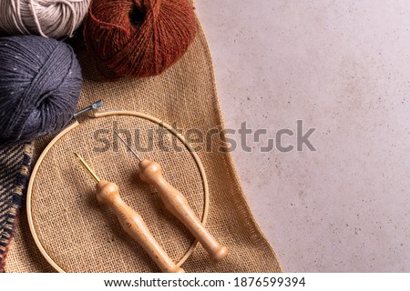 Composition with copy space of punch needle tools on a concrete surface. A jute fabric in an embroidery hoop, two different size of needles and three wool balls.