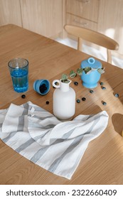composition of cotton dish towel, ceramic vase with eucalyptus branch, glass of water and fresh blueberry on wooden tabletop, high angle shot