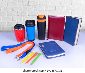 Composition Of Different Promo Products -Thermo Mugs, Lanyards Neck Strap, Pens, Notebooks For Business. On Desk Grey And Background White Wall