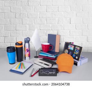 Composition Of Different Promo Products - Thermo Mug, Mug, Gifts, Pens In Boxes, Notebooks, Tools, Cap,flag Table. Copy Space. Grey Wall Background.