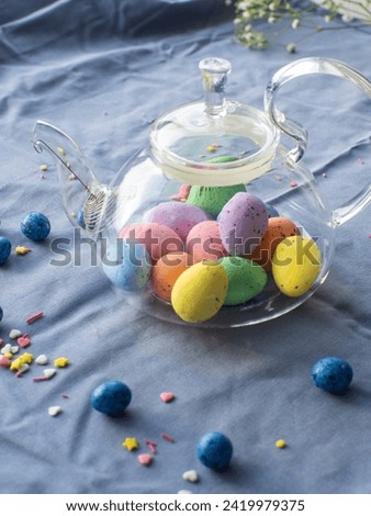 Composition with easter eggs in glass teapot surrounded by small chocolate eggs and sweet sprinkles. Easter tea concept. Tea pot full of colorful eggs on a blue background with copy space.