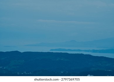 The composition emphasizes the calming qualities of misty, soft-focus horizons. The far-off island acts as a central visual point on the water, framing the relationship between untouched mountain peaks and the vast, geometric imprint of human civilization below.