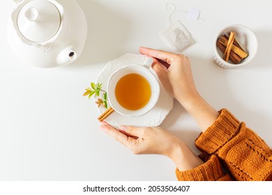 Composition with female hands holding a cup of tea, teapot, cinnamon and tea bag on white background. Flat lay, top view. Slow morning concept.