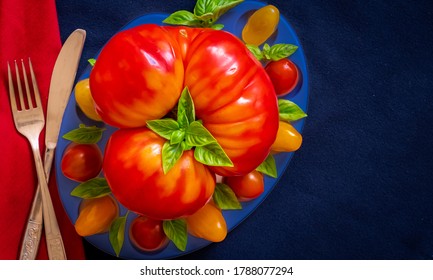 Composition with fork and knife on a red napkin and large heirloom tomato on blue tray surrounded with cherry tomatoes and decorated with basil leaves on dark blue background