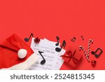 Composition with music sheets, note signs, Christmas gift and Santa hat on red background