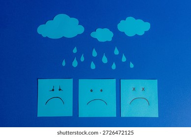 Composition on a blue background featuring three sad face sticky notes under paper cutout clouds and raindrops.