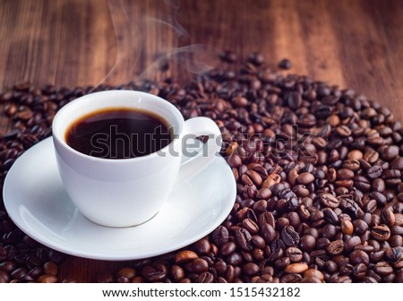 Composition of roasted coffee beans pilled round white cup full of aromatic espresso, close up view. Texture of brown wooden table. Selective soft focus. Blurred background