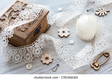 Composition with sewing tools: ivory thin lace, small light buttons, wooden flat flowers and safety pin as decor, wooden carved box and light candle wrapped in lace on the rustic surface. Flat lay