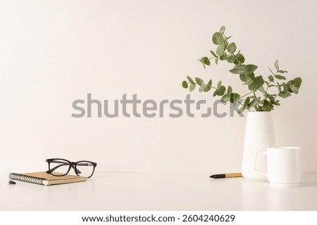 A composition showing a serene workspace setup with a notebook, glasses, eucalyptus, and white ceramic items on a white surface, providing a minimalist and tidy aesthetic