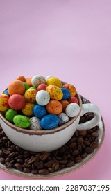 Composition with small chocolate easter eggs in cup surrounded by coffee roasted beans. Easter coffee concept. Mug full of colorful eggs on a plate with coffee beans on a pink background.