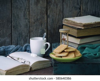 The composition of a stack of old books, open book, tea cups, glasses and plates of sugar cookies on a wooden background. Vintage photo