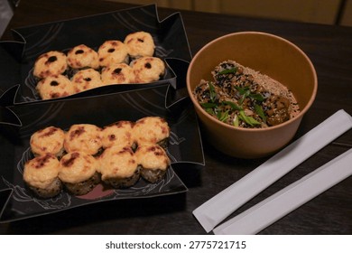 Composition of two black containers with baked sushi rolls, a brown bowl with rice and toppings, and two wrapped chopsticks. Shot on a dark wooden surface.