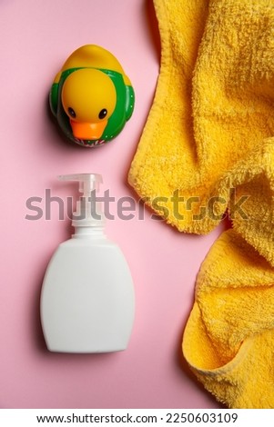 The composition of white clear liquid soap bottle with dispenser, bright yellow towel and multicolored child bathing duck on the light pink background. vertical view, baby bathing