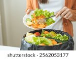 Compost from leftover food, asian young housekeeper woman hand holding plate use fork scraping waste, rotten vegetable throwing away into garbage, trash or bin. Environmentally responsible.